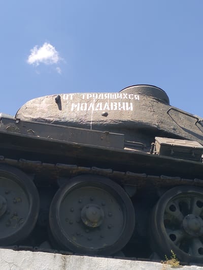 The image shows a close-up, low-angle view of the turret and tracks of a military tank, likely a monument, against a bright blue sky with a single small cloud. White Cyrillic lettering is painted on the turret. The text appears to read "ОТ ТРУДЯЩИХСЯ МОЛДАВИИ" which translates to "FROM THE WORKING PEOPLE OF MOLDOVA." The tank is dark green and shows signs of weathering. The tracks and road wheels are clearly visible. The tank is positioned on what appears to be a concrete base. The overall scene suggests an outdoor memorial or monument, likely located in the city of Cinișeuți, Moldova, given the inscription.