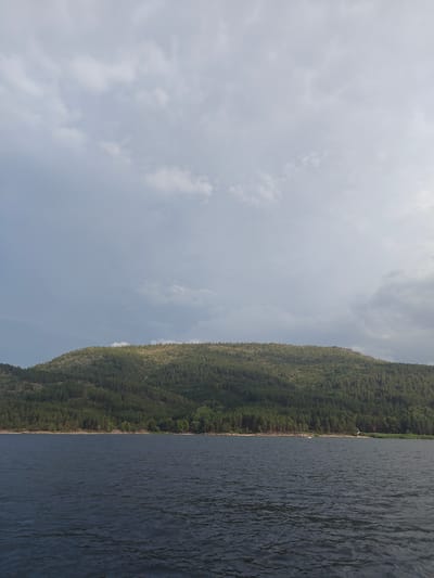 The image depicts a vast body of dark blue water in the foreground, with visible ripples indicating a light breeze. In the background, a densely forested hill rises, its green foliage stretching across the horizon. The top of the hill is lighter in color, possibly due to sparse vegetation or dry grass. Above the hill, a wide expanse of sky is filled with a mix of light gray and white clouds, with some areas appearing darker, suggesting overcast conditions or approaching rain. There are a few small structures visible along the shoreline at the base of the hill, including what appears to be a red-roofed building and a small white boat with people on it. The overall scene conveys a natural, somewhat somber landscape. The specific location in Manat, Kazakhstan, is not visually identifiable from the image alone.