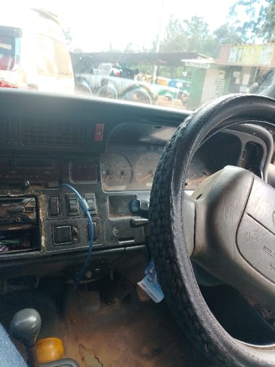 The image provides an interior view from a vehicle, looking outward through the front windshield onto a scene in Limuru, Kenya. In the foreground and midground, the vehicle's dashboard is visible; it is dark-colored, exhibiting signs of wear and dust. The dashboard includes a radio unit, various buttons, a prominent red hazard light switch, and an instrument cluster with visible gauges. A blue cable is connected to the dashboard area, hanging downwards. The steering wheel, positioned on the right side of the frame, is fitted with a textured black cover, and a blue banknote is tucked near its base on the light-colored steering column. To the lower left, a grey manual gear shift knob and a yellow object are partially visible, with the dusty floorboard and vehicle pedals discernible below.

Through the windshield, a bright outdoor environment under clear skies is observed. On the far left, a portion of a white van or bus is visible. Further into the background, multiple vehicles, including one with distinctive green and white markings, are present, some appearing to be under a corrugated metal roof structure. On the right side of the exterior view, a light-colored commercial building with a green-trimmed roof and unreadable signage is visible, consistent with a small shop or kiosk. Trees and other vegetation are visible in the distant background. No individuals are clearly identifiable in the scene.