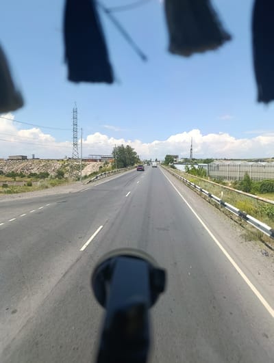The image captures a view from a vehicle traveling on a highway in Dzoraghbyur, Armenia. The road is asphalt, with clear lane markings and a guardrail lining the right side. The surrounding environment features dry, rocky terrain with sparse vegetation. In the distance, several structures are visible, including industrial buildings, a tall power pylon, and what appear to be greenhouses. The sky is blue with scattered white clouds, suggesting fair weather. Several other vehicles are also present on the highway, moving in the same direction as the viewpoint vehicle. The foreground is partially obscured by a dark, blurry object, likely part of the vehicle from which the photo was taken.