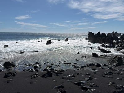 The image shows a black sand beach in Yaiza, Spain. The foreground is filled with black sand and rocks. The ocean waves are crashing onto the shore. The sky is blue with some clouds.