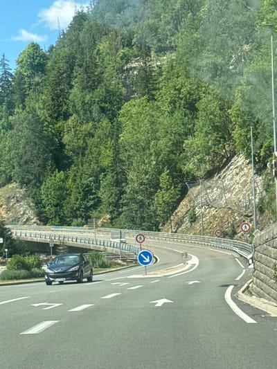 The image captures a road in the city of Hauts de Bienne, France, likely near a mountainous area given the dense evergreen forest covering the hills. The road is paved and marked with white arrows indicating lane directions. In the foreground, a dark colored hatchback car is visible, driving on the road. A concrete bridge spans across the road in the middle ground. Traffic signs are present, including a speed limit sign indicating 70, a pedestrian crossing sign, and a blue circular sign with a white arrow indicating a mandatory turn. The environment appears to be a sunny day with a clear blue sky and some scattered clouds. The road curves to the right, suggesting a winding mountain pass. A stone wall is visible on the right side of the road, and a metal guardrail runs along the edge of the road and the bridge.
