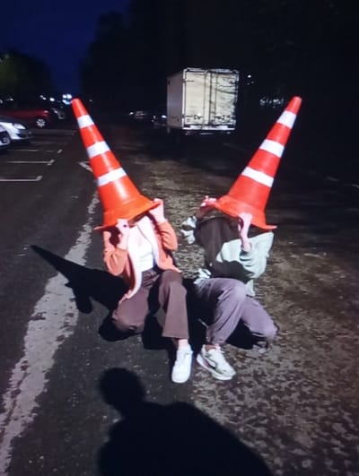 Two individuals are depicted at night in Kukcha, Uzbekistan, crouching on a dark, wet-looking paved surface. Each person has an orange traffic cone with two white reflective stripes positioned over their head, obscuring their faces. The individual on the left wears a light-colored top beneath an orange jacket and brown pants, with white sneakers. The individual on the right wears a light-colored long-sleeved top and purple pants, with light-colored sneakers. Both subjects are holding the cones over their heads with one hand. Long shadows extend from their figures towards the foreground. In the background to the left, multiple parked vehicles are visible in a designated parking area. To the center-right, a white box truck with a dark trailer is present. The scene is dimly lit, with the foreground subjects appearing illuminated by a direct light source.