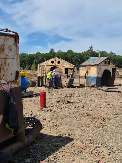 In a construction site in Taezhnoe, Russia, two individuals in work attire are engaged in an activity around a large industrial valve and piping system. The scene is an outdoor, rocky terrain with scattered debris, suggesting ongoing construction or demolition work.

The foreground is dominated by a large, rusted metal container on the left, partially obscuring a bright blue industrial valve with yellow fittings. A red cylindrical object, possibly a fire extinguisher or a fuel can, stands upright on the ground nearby.

In the midground, two figures are visible. One, wearing a high-visibility yellow vest and white helmet, is standing and looking down. The other, wearing a blue overall and white helmet, is bent over, possibly working on the equipment. Behind them are two dilapidated, box-like structures made of rusted metal, resembling makeshift shelters or equipment housings. One of these structures has a ladder extending upwards.

The background features a dense line of green trees under a partially cloudy blue sky. The ground is uneven and covered with gravel and dirt, with an orange cable snaking across the surface. Several metal pipes are also scattered around the site. The overall impression is one of an industrial work zone in a rural or semi-rural setting.