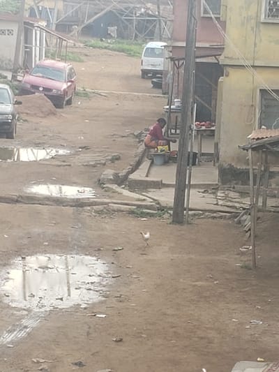 The image shows a street scene in Alalubosa, Nigeria. The road is unpaved and muddy with puddles reflecting the sky. A woman wearing a red top and patterned skirt is bending over a bucket near a building. A small wooden structure with displayed goods sits next to her. Vehicles, including a white van and a red car, are parked along the side of the road. Buildings of varying construction are present, some with wooden frames and others with more substantial walls. Power lines stretch across the scene.