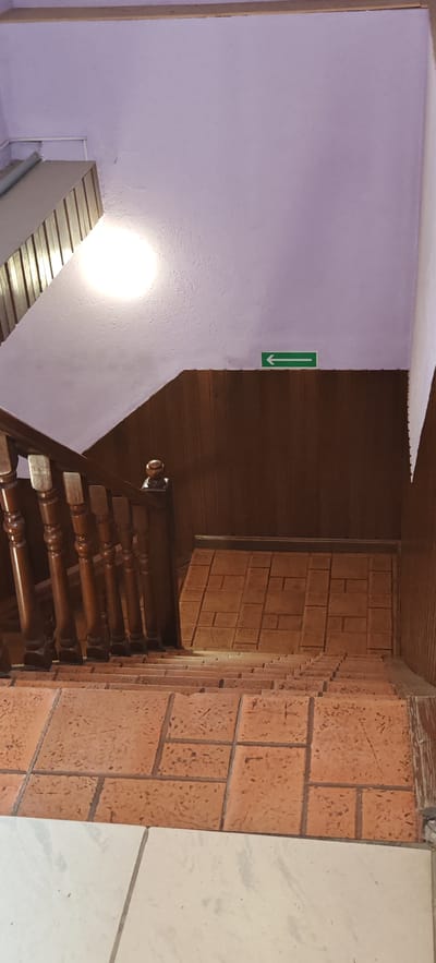 The image shows a staircase from a top-down perspective. The walls are painted a light purple, and the lower portion of the wall is paneled with dark wood. There is an illuminated green arrow sign pointing to the left on the purple wall. The staircase itself has wooden banisters with turned spindles and a wooden handrail. The steps are covered with terracotta-colored tiles, and there is a landing with similar tiling. The floor at the bottom of the image is covered with light-colored, possibly marble, tiles. The scene appears to be indoors, possibly in a residential building or a public space. No people are visible in the image. The lighting suggests an artificial light source, possibly a ceiling light that is out of frame. The environment seems clean and well-maintained.