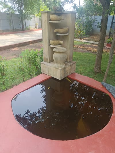 The image depicts a water feature in Dhundshi, India, likely a garden or backyard setting. The feature consists of a tiered stone structure with bowl-shaped elements and a large vase-like component at the base, positioned above a small, irregular-shaped pond. The pond is bordered by a reddish-brown concrete surface, and the water within appears dark and reflective, showing the reflection of foliage from surrounding trees. The area around the water feature includes grass, trees, and various plants, with a concrete or paved pathway and a low concrete wall visible in the background. A gray cloth or covering lies near the edge of the pond. The overall scene is outdoors and appears to be in a residential or recreational area.