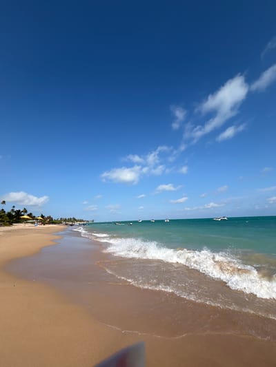 The image displays a sunlit beach scene in Maragogi, Brazil. The upper two-thirds of the frame consists of a clear blue sky with sparse, scattered white cumulus clouds. The lower third features the ocean and a sandy beach. The ocean exhibits a turquoise coloration, with white-capped waves actively breaking onto the shore in the midground. Multiple boats and personal watercraft are visible in the distant ocean. The beach consists of light brown sand, stretching into the distance along the coastline. On the far-left shoreline, several palm trees, yellow beach umbrellas, and numerous indistinct human figures are visible on the sand and in the shallow water, indicating recreational activity.