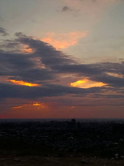 This image captures a sunset over the city of Orto-Sai, Kyrgyzstan. The sky is a canvas of dramatic clouds, with hues of orange, pink, and gray. Sunlight pierces through a gap in the darker clouds, casting a bright yellow glow. Below, the cityscape is silhouetted against the fading light, with buildings and structures visible in the distance. The foreground is a dark, hilly terrain with some rocks and vegetation, suggesting a viewpoint from a higher elevation overlooking the city. The overall atmosphere is one of natural beauty and urban expanse.