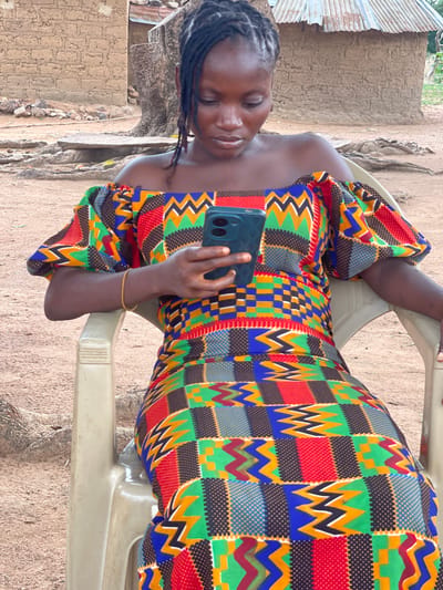 A young Black woman is depicted in an outdoor setting, seated and engrossed in her smartphone. Her hair is styled in short dreadlocks or twists, and she has a calm, focused expression as she looks down at the device. She is wearing a vibrant, floor-length dress adorned with a complex, multi-colored geometric pattern, typical of West African textiles, featuring squares, zigzags, and dots in hues of red, green, blue, yellow, and black. A thin, yellow bracelet is visible on her right wrist.
The woman holds a dark-colored smartphone with her right hand, showcasing a distinct triple-camera module on its back. She is seated on a light-colored, possibly beige or off-white, plastic chair.
The background reveals a simple, earthy environment, consistent with a rural or semi-urban area in Tabu, Nigeria. To her left, a structure with corrugated metal sheeting (likely a roof or wall) stands beside a textured wall made of mud or plaster. The ground beneath her and extending into the background is dry, earthy soil with visible small rocks and roots. The bright, even lighting suggests daytime, likely mid-morning or afternoon, under clear weather conditions. The scene captures a moment of modern technology being used within a traditional setting. No discernible text is visible within the image.