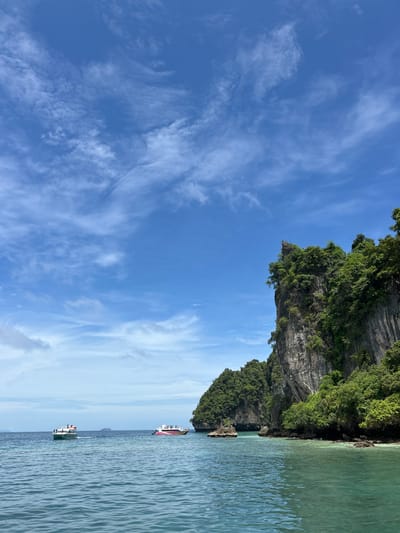The image depicts a scenic view of islands and the ocean in Ko, Thailand. In the foreground, the clear turquoise water shows gentle ripples. In the middle ground, two boats are visible on the water. The boat on the left is green and white with several people on deck. The boat on the right is white with a colorful striped hull, also carrying passengers. In the background, a large, rocky island covered in lush green vegetation rises dramatically from the sea. The sky above is a vibrant blue, dotted with wispy white clouds, suggesting a clear, sunny day.
