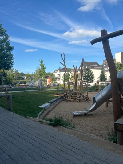 The image depicts a children's playground in Waldbröl, Germany, on a clear day. The scene is dominated by a modern, nature-inspired play structure constructed primarily from wood and featuring a metal slide. The playground has a sandy surface area and is bordered by a green lawn. In the background, there are residential buildings and trees, suggesting an urban or suburban setting. The sky is a vibrant blue with scattered wispy clouds, indicating fair weather and likely daytime. There are no people visible in the image, implying the playground is either empty or the image was taken at a quiet moment. The overall impression is one of a well-maintained and safe outdoor play area designed to blend with its natural surroundings.