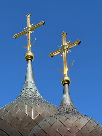 This low-angle, medium shot captures two identical, ornate golden Orthodox crosses atop the conical, weathered copper domes of a church. The sky is a vibrant, clear blue. Each cross features a sunburst design at its center and a crescent moon below. The domes are covered in diamond-shaped copper tiles, showing signs of age and patina. The scene is a static, architectural composition devoid of any people or discernible activity, suggesting a focus on the religious structures against the backdrop of the sky. The location is identified as St. Ozerki, Russia.