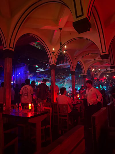 The image shows the interior of a dimly lit bar or club. The vaulted ceiling has decorative arches. Speakers are mounted high on the walls. People are seated at tables, and there are multiple glasses and bottles on the tables. The scene is lit with red and blue lighting, creating a nightclub atmosphere. The exact location in Xuanwu District, China, cannot be determined with certainty.