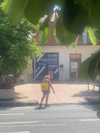 In the city of Stupava, Slovakia, on a sunny day, a person is seen from behind, wearing a bright orange vest and a yellow backpack sprayer. They are on a paved sidewalk, which transitions to a road with white lane markings. The person is holding a long wand and appears to be spraying something onto the ground or vegetation near the sidewalk's edge, possibly for pest control or maintenance. Behind the person, a building with white siding and a tiled roof is visible. A large blue sign on the building features text in Slovak and a circular red and white "no parking" symbol. To the left of the building entrance, a tree with lush green leaves shades a small outdoor seating area with a white folding table and two chairs. A large planter filled with dark red flowers is situated near the seating area. To the right of the main building entrance, there is another doorway labeled "NECHTOVE STUDIO BEAUTY SALON" with a small sign advertising services. The overall scene depicts a street-level view of an urban setting with a person engaged in outdoor maintenance.