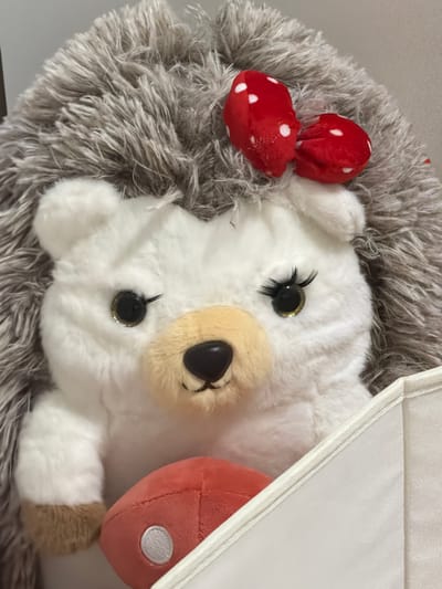 This media file features a close-up view of a plush toy hedgehog. The hedgehog is predominantly white with gray, furry spines. It has black, glittery eyes with long lashes, and a light brown snout. Two red bows with white polka dots are attached to the top of its head, positioned like ears. A small, red, mushroom-shaped object with white spots is also visible near the hedgehog's head. Part of a white, structured object, possibly a box or container, is visible in the upper right corner, casting a shadow. The plush toy is resting on a textured surface, also gray and furry. The lighting suggests an indoor setting, with no specific time of day or weather conditions discernible. No text is visible in the image. The overall impression is of a cute and whimsical object, likely a toy intended for children.