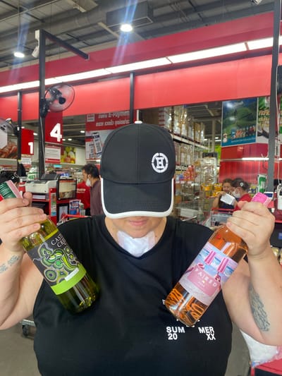 The image displays a person holding two bottles of wine inside a brightly lit retail store, likely a supermarket or wholesale club. The primary subject, seen from the chest up, wears a black t-shirt with "SUM 20" and "MER XX" text, a black baseball cap featuring a white, stylized logo, and a white bandage on their neck/face. Tattoos are visible on their arms.

They hold a pink bottle of "PAIRED ROSÉ Wine of Australia" with a label depicting popsicles on a beach, and a green-labeled bottle of "CASTLE CREEK" white wine, which features a distinctive green, black, and white indigenous-style art pattern.

The background shows a typical store environment with multiple checkout counters, one marked with a large "4," and shelves stocked with various products. Other shoppers and staff, some wearing face masks, are visible in the aisles. Overhead strip lighting and ceiling fans illuminate the space. Visible text on store signs includes "E-Payment" and Thai script reading "Convenient, fast, all payment channels." A "Dilmah" tea poster with a landscape and the word "makro" are also discernible, confirming the setting as a Makro store in Moo 1, Thailand. The activity appears to be shopping, with the individual holding the wine bottles, possibly at a checkout.