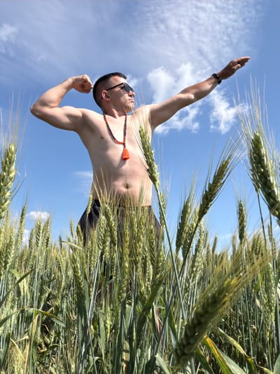 A shirtless man wearing sunglasses and a beaded necklace with an orange tassel poses in a wheat field under a blue sky with scattered clouds. His left arm is flexed at the elbow, and his right arm is extended horizontally to his right, with his hand in a flat, open position. The man appears to be standing in a field of mature wheat, with the stalks of wheat prominent in the foreground and surrounding him. The sky is bright and clear, indicating daytime. The location in Podshivalovo, Russia is not discernible from the image itself.