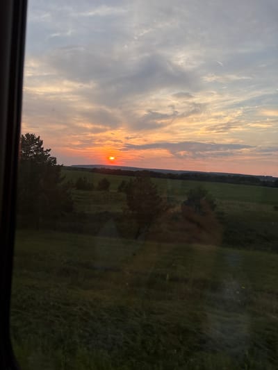 The image shows a sunset over a rural landscape in the city of Beryozovka, Russia. The sun, a bright orange orb, is low on the horizon, casting a warm glow across the sky, which is filled with streaks of orange, pink, and grey clouds. In the foreground, there are dark silhouettes of trees and rolling green hills. The overall scene is peaceful and serene, with the setting sun creating a beautiful and dramatic atmosphere.