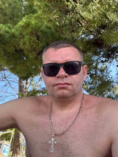 A man with short graying hair, wearing black sunglasses and a gold chain with a cross pendant, poses for a selfie outdoors. He is shirtless, revealing his chest and shoulders, with a pine tree and blue sky visible in the background. The scene appears to be a beach or a park in Makarska, Croatia.