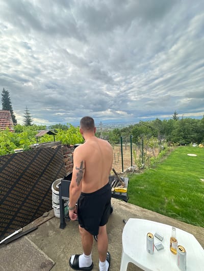 The image captures a shirtless man in black shorts and black and white slide sandals, standing outdoors next to a grill. He has a tattoo on his left arm. The environment features a view of a city in the distance, nestled among trees and greenery. Adjacent to the man is a surface with the words "SKYFORM" printed repeatedly on it. Further in the background is a table holding beverage cans and a bottle. The setting appears to be in Brestnik, Bulgaria. The sky is overcast with dense clouds.