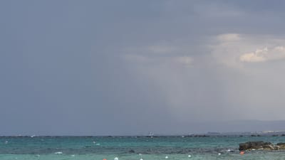 The image depicts a cloudy day over a body of water, likely the sea, with a distant shoreline visible. The water is a turquoise color and appears choppy with whitecaps. Scattered across the water are several white and orange buoys, and some rocks are visible along the coastline and protruding from the water. In the distance, a ship is sailing. The sky is filled with dark, heavy clouds, suggesting impending rain or a storm. The overall atmosphere is somber and overcast. This scene is not identifiable as being in Taranto, Italy.