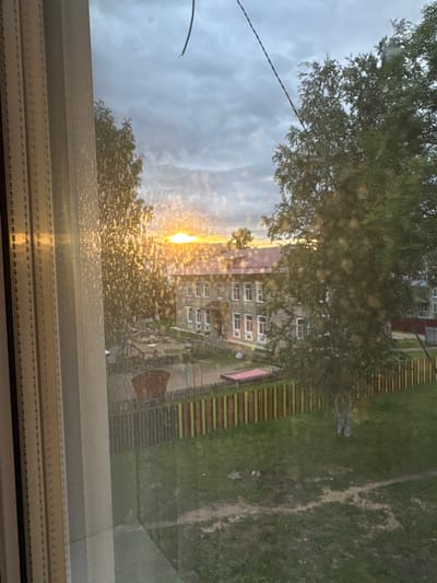 A view through a window shows a school building in the city of Kuvshinovo, Russia, during sunset. The sun is low on the horizon, casting a warm glow across the sky and illuminating the leaves of trees in the foreground. The school is a multi-story brick building with a red roof. A fence with colorful vertical slats surrounds the schoolyard, which contains playground equipment and grassy areas. The window has a curtain on the left side and a thin wire or string running across the upper portion of the image. The overall scene is serene, with the soft light of sunset creating a peaceful atmosphere.