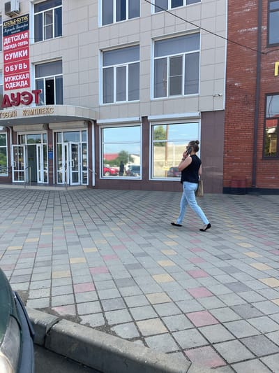 A person is standing on a sidewalk in front of a building, looking at their phone. They are wearing a black t-shirt and light-colored jeans. The building appears to be a commercial establishment with large windows and a sign that reads "ДУЭТ ТОВЫЙ КОМПЛЕКС" in Russian, which translates to "DUET SHOPPING COMPLEX." Below this, there are signs for "АТЕЛЬЕ-БУТИК," "ДЕТСКАЯ ОДЕЖДА ОБУВЬ," "СУМКИ," and "МУЖСКАЯ ОДЕЖДА," indicating various retail shops within the complex. The sidewalk is paved with colorful square tiles. The time of day appears to be daytime, with clear sunlight casting shadows. The overall scene suggests a typical street view in a town, possibly during pleasant weather.