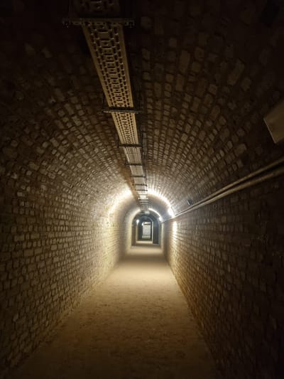 This is an underground tunnel in Homburg, Germany. The tunnel is constructed from tan-colored bricks that form an arched ceiling, floor, and walls. A metal railing runs along one side of the tunnel. Light fixtures are attached to the ceiling, providing illumination. The tunnel narrows in the distance, leading to a rectangular opening at the far end. The far end appears to be brighter than the immediate foreground.