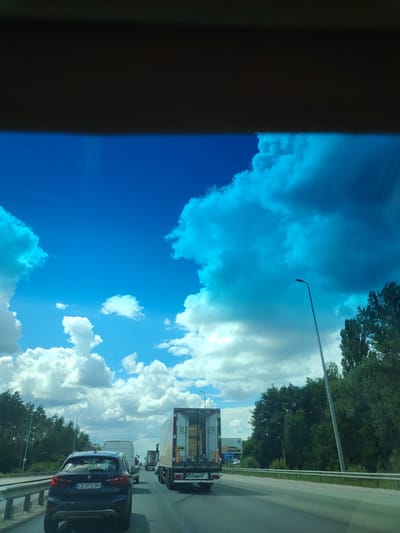 A forward-facing, eye-level shot captures traffic on Peremohy Street in Ukraine. The scene is dominated by a bright blue sky filled with dramatic, white and turquoise clouds. In the foreground, a dark blue BMW SUV with a license plate reading "CB 2012 BM" is in the left lane. To the right, a white truck with "ROS HEN" branding is in the right lane. Behind it, other vehicles and trees line the road, and a tall lamppost stands beside the highway. The road appears to be a multi-lane highway with a metal guardrail separating the lanes. The overall impression is of a sunny day with heavy clouds, likely during daytime hours.