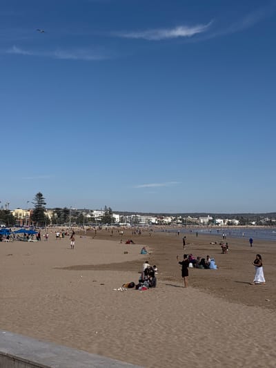 A wide, sandy beach in Essaouira, Morocco, is populated with numerous people enjoying the day under a clear blue sky. In the foreground, a concrete barrier separates the sand from a lower paved area. The beach stretches towards a distant coastline lined with buildings and palm trees, indicating an urban area. People are scattered across the sand, some sitting in groups, others walking, and a few are in the shallow water in the background. Beach umbrellas and loungers are visible towards the left side of the frame, suggesting a developed beach area. A lone bird flies in the upper left quadrant of the sky.