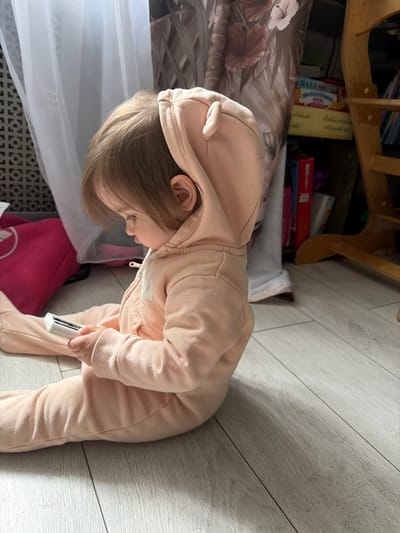A young child wearing a pink hooded jumpsuit stands on a light gray wooden floor holding a white rectangular object with a black element. The child is bending over and appears to be looking at something near the floor. Behind the child are portions of furniture and items, including a light-colored wooden frame with a shelf, a patterned surface resembling bedding, and a pink object. The location cannot be confirmed as Novosergievka, Russia, based on the image content.