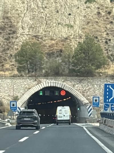 The image captures a view of a highway leading towards a tunnel entrance in Lorca, Spain. Two vehicles, a dark SUV in the foreground and a white van in the middle lane, are proceeding towards the tunnel. The tunnel entrance is illuminated by lights and features traffic signals and signage. Signs indicate "80" (likely a speed limit) and "Lorza 1000 m" (indicating Lorca is 1000 meters away). The tunnel is constructed with an arched stone facade. The surrounding environment is dry and hilly, with sparse vegetation. The road has visible lane markings and barriers along the side. The lighting suggests it is daytime.