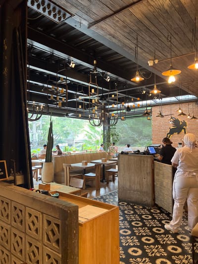 The photograph depicts a cafe interior. A wooden ceiling holds industrial-style light fixtures. Large windows offer views of lush greenery. Seating includes cushioned benches and wooden chairs. A geometric tiled floor contrasts with the rough concrete and wood elements of the counter. Staff dressed in white uniforms are visible. The composition emphasizes the cafe's design, blending natural light and modern aesthetics.