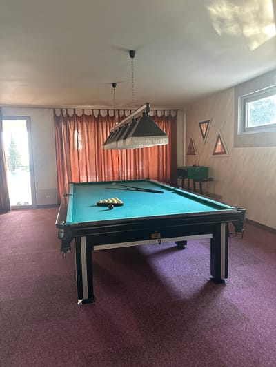 The image depicts a room in a private residence or establishment, likely a game room, located in Brca, Montenegro. The central object is a green pool table with black trim and legs, positioned on a purple carpeted floor. Several billiard balls, including a red one and a rack of white balls, are placed on the table. Above the pool table, a multi-lamp lighting fixture hangs from the ceiling. In the background, there are large, rust-colored curtains covering a window and a glass door that leads outside to a bright area with trees and a railing. The walls are painted in a light, textured color, with several triangular decorative frames on one wall. To the right, a small green cabinet sits on a stand near a window with a view of trees. The room is illuminated by natural light entering from the door and window, as well as the overhead lamps.