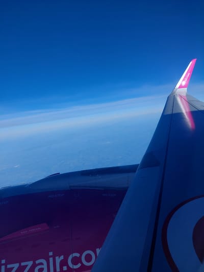The image shows the wing of a Wizz Air airplane in flight, seen from a passenger window. The wingtip is painted with the Wizz Air logo in pink and white. The aircraft is flying over a hazy blue sky. The location of the city of Pyrzowice, Poland is not discernible from the image.