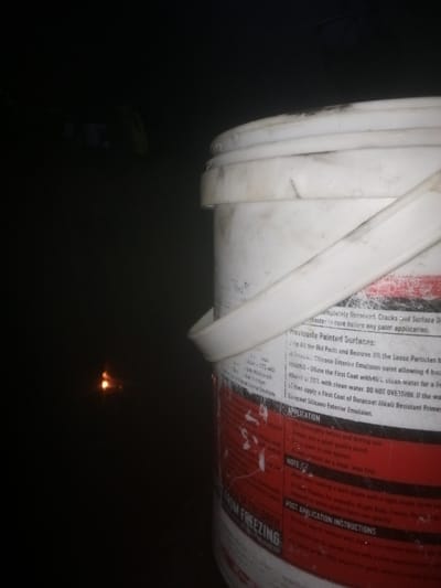 A partially visible, white paint bucket with a plastic handle sits in the dark. The bucket's label contains usage instructions, including surface preparation, application, and post-application steps. It warns against dilution overestimation and suggests using a Duracoat Alkali Resistant Primer. The text also cautions to keep the paint from freezing. Light glints off the label, making the text more legible. The bucket is dirty and worn.