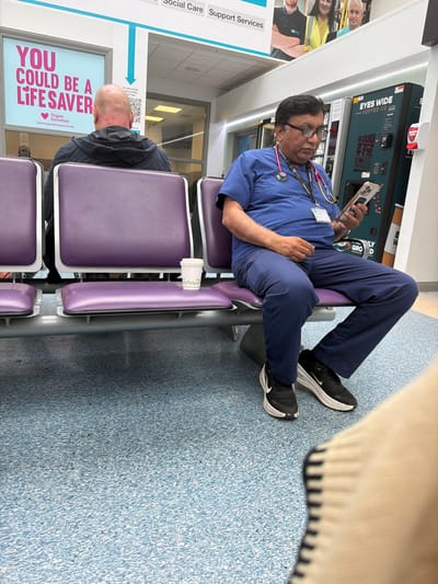 The image depicts a waiting area, likely within a hospital or medical facility in Portadown, United Kingdom. The primary subject is a man of South Asian descent, wearing blue medical scrubs, glasses, and a stethoscope around his neck. He is seated on a purple waiting room chair, with his legs propped up on an adjacent chair, engrossed in his smartphone. His identification badge clearly visible on his chest states "Consultant" multiple times. He wears black and white Nike sneakers.

To his right, another man with a bald head and wearing a dark top is seated on a similar purple chair, facing away from the camera. The floor is a light blue speckled pattern.

In the background, a "EYES WIDE COFFEE CO." vending machine is visible, displaying "2.30" among other numbers, indicating a price. Adjacent to it, a large poster encourages "YOU COULD BE A LIFESAVER" and "Organ Donation" with the website "www.organdonationni.info". Another sign mentions "Social Care Support Services" and "To view all job opportunities, scan here" next to a QR code. A white paper cup rests on one of the empty purple chairs. The scene suggests a period of rest or waiting in a public institutional setting.