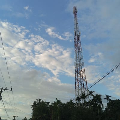 The image captures a telecommunications tower under a partly cloudy blue sky in Uleejalan, Indonesia. The dominant subject is a tall, lattice-structured tower, painted in alternating red and white sections, positioned vertically on the right side of the frame. Multiple antenna arrays and equipment boxes are mounted on its upper and middle sections. In the foreground, spanning the bottom of the image and extending partially up the left side, is a dense canopy of tropical trees, including several tall palm trees with visible fronds. Power lines or utility cables stretch diagonally across the upper left and center-right of the image, with a utility pole partially visible on the far left edge, equipped with insulators. No people or specific actions are discernible. The environment appears to be an outdoor setting, likely a rural or suburban area given the vegetation and utility infrastructure.