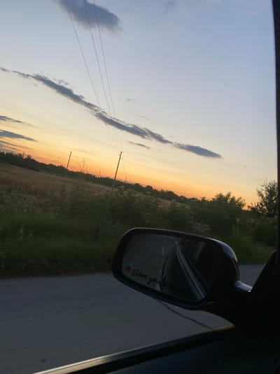 The image depicts a view from a car window in Miriantsi, Bulgaria, during sunset. The sky is a gradient of pale blue to orange, with wispy clouds and power lines visible. A field with tall grass and some trees lines the roadside. The side mirror of the car is visible, reflecting the road ahead and displaying a small red heart with the words "I love you" written below it.