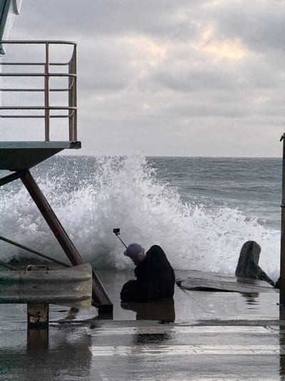 The image shows a person kneeling on a wet pier, holding a selfie stick with a camera attached. The person is wearing a dark hooded jacket and a purple cap. Behind the person, a large wave crashes against the pier, sending white spray into the air. In the background, the ocean stretches to the horizon under a cloudy sky. To the left of the frame, a metal railing and part of a structure are visible, possibly a lifeguard stand or a pier amenity. To the right, a dark, weathered piece of wood or debris is partially submerged in the water. The pier itself appears to be made of concrete and wood, with puddles of water reflecting the overcast sky. The scene captures a moment of dramatic natural activity near the coast, with the person seemingly focused on capturing the event. The specific location is not explicitly identifiable from the image alone, but it depicts a common scene found along coastal cities in the United States of America.