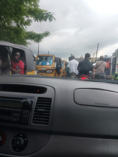 The media file depicts a street scene in Iju, Nigeria, likely during the daytime with overcast skies. The perspective is from inside a vehicle, looking out through the windshield at traffic.
The foreground is dominated by the interior of the vehicle's dashboard, showing a radio with a digital display, ventilation grilles, and control knobs. A text label "SRS AIRBAG" is visible on the dashboard.
Outside the vehicle, a variety of modes of transportation and people are visible. Immediately to the left, a covered auto-rickshaw, known locally as a "Keke Napep", carries a man wearing a red t-shirt. To the right of this, a bright yellow "Keke Napep" with a license plate reading "JND 6392" is stopped in traffic. Beyond this, several individuals are seen on motorcycles. One man in a white shirt is on a motorcycle, and another in a black shirt is also riding a motorcycle, with his brake light illuminated, suggesting he may be stopped or slowing down. Further in the background, more people, motorcycles, and what appears to be a truck or larger vehicle can be seen. Some individuals are wearing high-visibility vests, indicating they may be involved in traffic management or construction.
The overall atmosphere appears to be that of a busy, congested road. The presence of multiple "Keke Napep" vehicles and motorcycles is characteristic of traffic in many Nigerian cities. The cloudy weather suggests a potentially humid or rainy day, common in tropical climates. The scene captures a moment of typical urban transportation and daily life in Nigeria.
