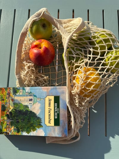 A still life image shows a woven string bag filled with green apples and a lemon. A red and yellow nectarine rests on top of the bag. A book with a painted cover depicting a church and a tree lies next to the bag. The objects are placed on a teal-colored wooden surface, possibly a table. The lighting suggests it is daytime, with shadows cast by the objects. The image does not provide any information about the city of Starokorsunskaya, Russia.