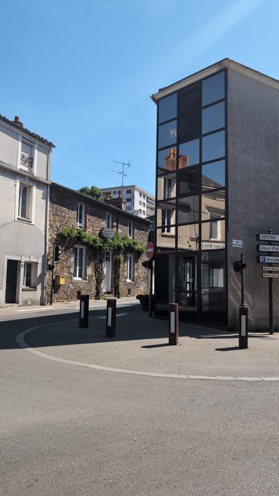 The image depicts a street scene in Cholet, France, likely at an intersection. On the right is a modern building with a facade of large, dark-tinted glass panels, reflecting the surrounding environment, including a stone building with white-framed windows and a chimney. A street signpost on this building indicates directions to "CENTRE", "Sous-Préf.", "P Arcade", "Hôtel Mercu.", and "Hôtel Ibis B.". Adjacent to this modern building, to the left, is a traditional stone building with green foliage climbing its facade, white-trimmed windows, and a white door. Above this stone building, a taller, multi-story building with white balconies is visible, along with antennas on its roof. A grey, two-story building with a white-trimmed window on the upper floor and a dark door on the lower floor stands to the far left. The street is paved with asphalt and a circular grey area with white lines. Several bollards are positioned in the foreground. The sky is clear and blue. No people are visible in the frame.