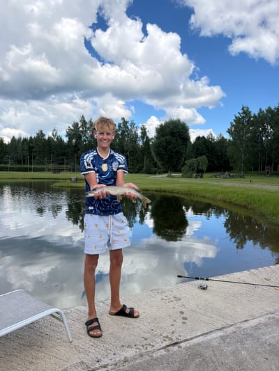A young man stands on a concrete platform next to a body of water, holding a freshly caught fish. He is wearing a blue and white soccer jersey and striped shorts with small yellow duck figures. He has blonde hair and is wearing white earbuds. To his right, a fishing rod with a reel rests on the concrete. The water reflects the cloudy blue sky and the surrounding trees. In the background, there is a grassy area with trees and some structures, suggesting a park or recreational area. The location is identified as Mežpļavas, Latvia.