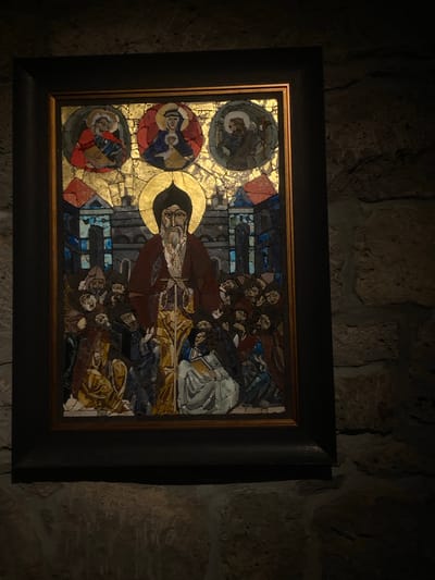 A framed mosaic artwork depicting religious figures is displayed on a stone wall. The artwork features three circular medallions on the left side and a central figure with a golden halo extending horizontally across the mosaic. Several other figures are arranged around the central figure, creating a busy composition. The mosaic consists of small, irregularly shaped pieces of stone or glass in various colors. The frame is dark and contrasts with the lighter mosaic. The wall is made of rough-hewn stones and the scene appears to be located in Tatev, Armenia.