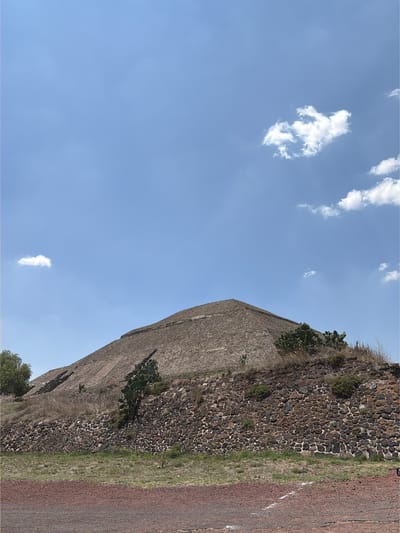 The image shows a portion of the Pyramid of the Sun in Teotihuacan, located in San Sebastián Xolalpa, Mexico. The pyramid is constructed of stone and earth, with some vegetation growing on its surface. To the right of the pyramid, there is a path or roadway. The sky is blue with scattered clouds. The image appears to have been taken during daylight hours.