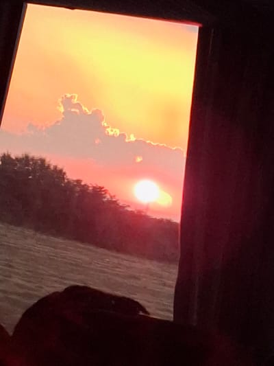 The image shows a sunset viewed through a window or frame, with the sky ablaze in vibrant oranges and yellows. A bright sun, partially obscured by clouds, is the focal point of the sky. The clouds themselves are illuminated by the setting sun, creating a dramatic silhouette against the fiery backdrop. To the right of the frame, a dark, indistinct shape suggests a silhouette of a tree or other vegetation, adding depth to the scene. The overall impression is one of a beautiful and serene sunset. The location is identified as Skutare, Bulgaria. There is no visible text in the image.