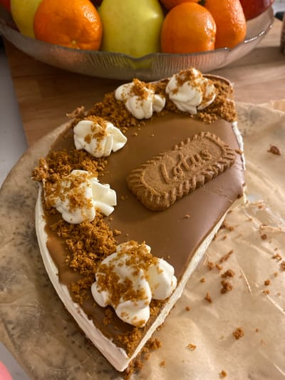A piece of Lotus Biscoff cheesecake rests on parchment paper. The cake has a smooth, brown caramel-colored top with a Lotus cookie positioned in the center. Whipped cream dollops are arranged along the curved edge, sprinkled with crushed Biscoff crumbs. The base appears to be made of white cream cheese filling. Adjacent to the cake is a glass bowl filled with tangerines, an apple, and other fruits. The background is a wooden surface.