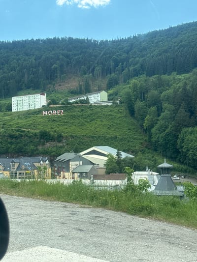 The image shows a hillside with the word "MOREZ" spelled out in large, red and white letters on the grassy slope. Two multi-story white buildings are situated higher up on the hill, one with a red roof and balconies, and another with a white roof. Below the large letters, several buildings are visible, including structures with multi-pitched roofs and a building with a distinctive grey, domed cupola. The overall setting is mountainous and forested, with a cloudy blue sky overhead. The scene appears to be in the town of Morez, which is now part of the city of Hauts de Bienne in France.